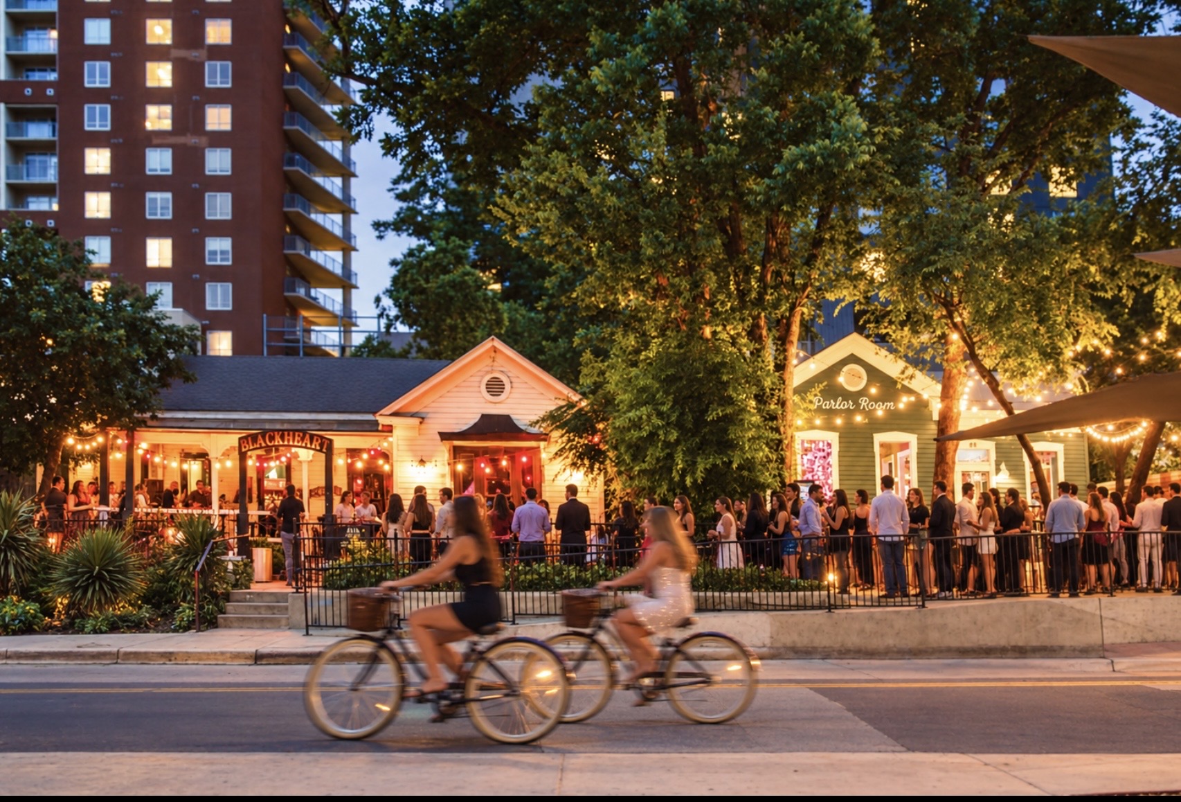 Rainey Street at dusk, with two bicyclists passing bungalow bars and a line of people enjoying the evening